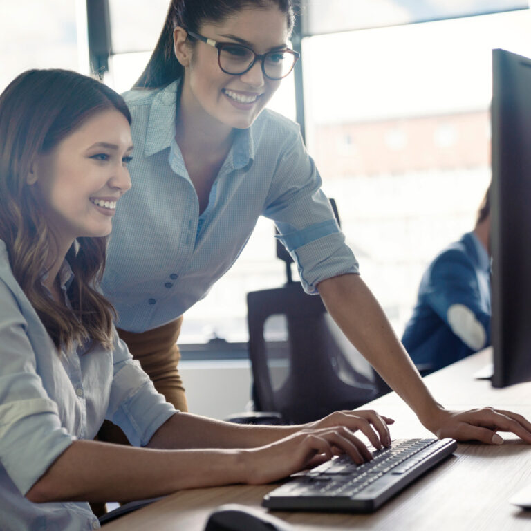 Zwei Frauen stehen bzw. sitzen vor dem Computer im Büro